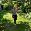White woman in her 50s with short hair and glasses, sitting on a swing in an overgrown lush green ga